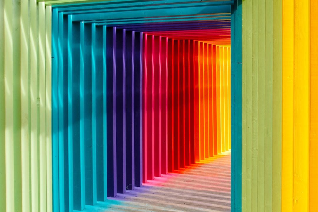 A colorful walkway with vertical, rainbow-colored panels casting shadows, creating a bright tunnel effect.