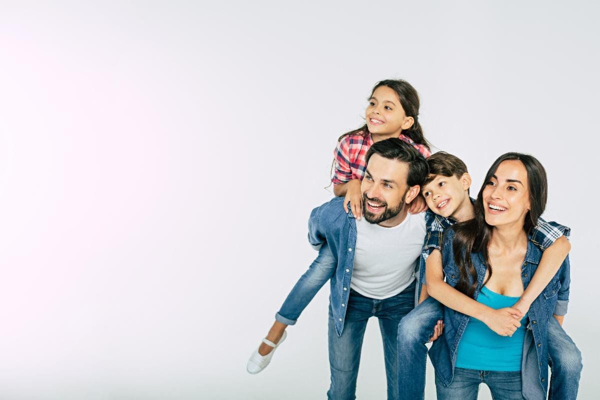 Family smiling, two children on parents' backs against a white backdrop.
