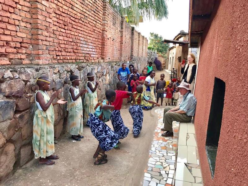 Children in patterned clothing dance in a narrow outdoor corridor while two adults watch from a wall.