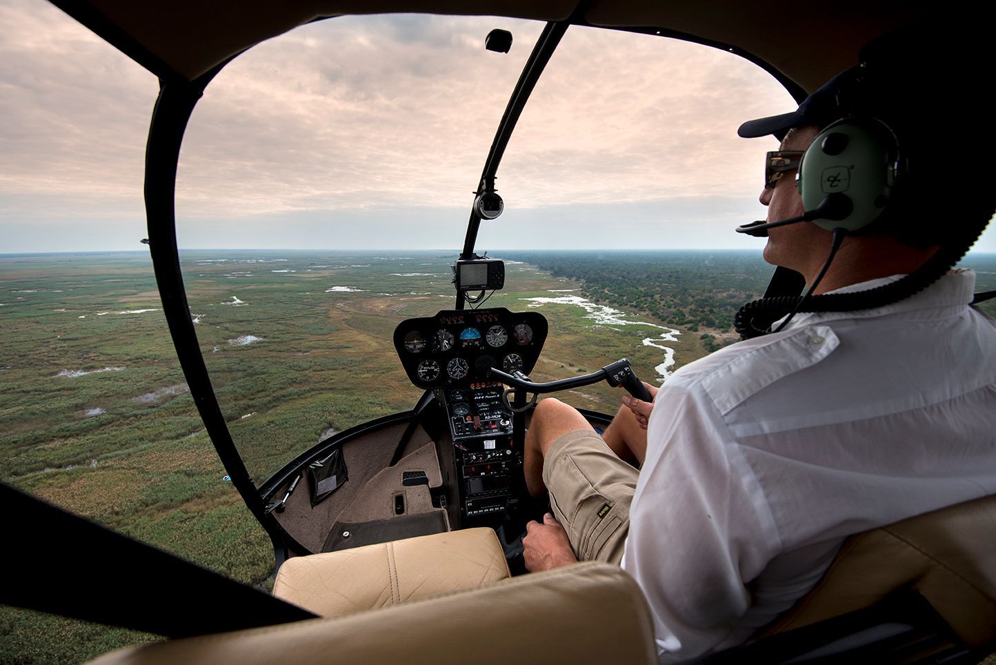 Pilot in the cockpit of a helicopter flying over a vast, green landscape with a winding river under a cloudy sky.