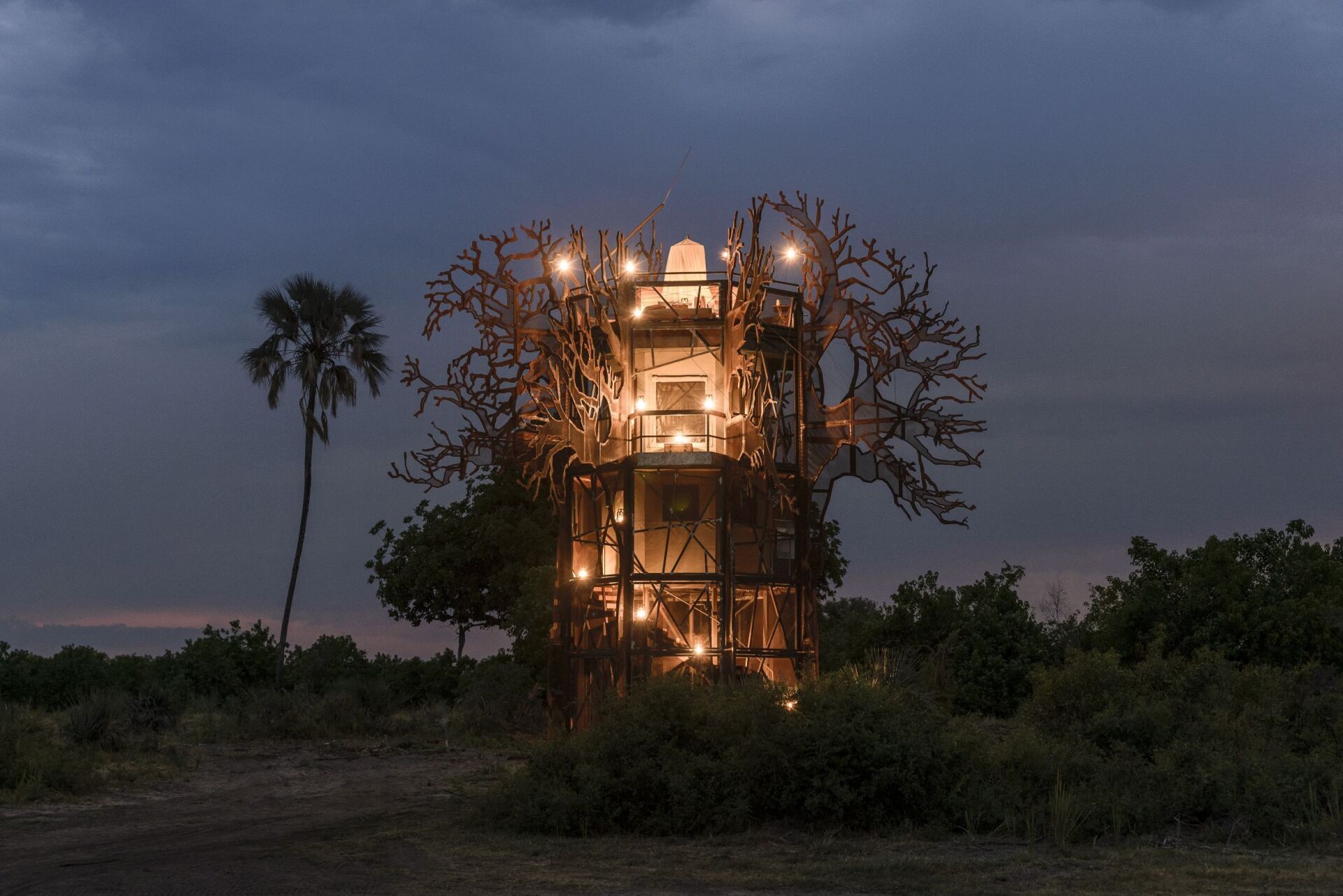 A multi-level wooden structure illuminated with warm lights at dusk, set against a dark sky in a grassy landscape.