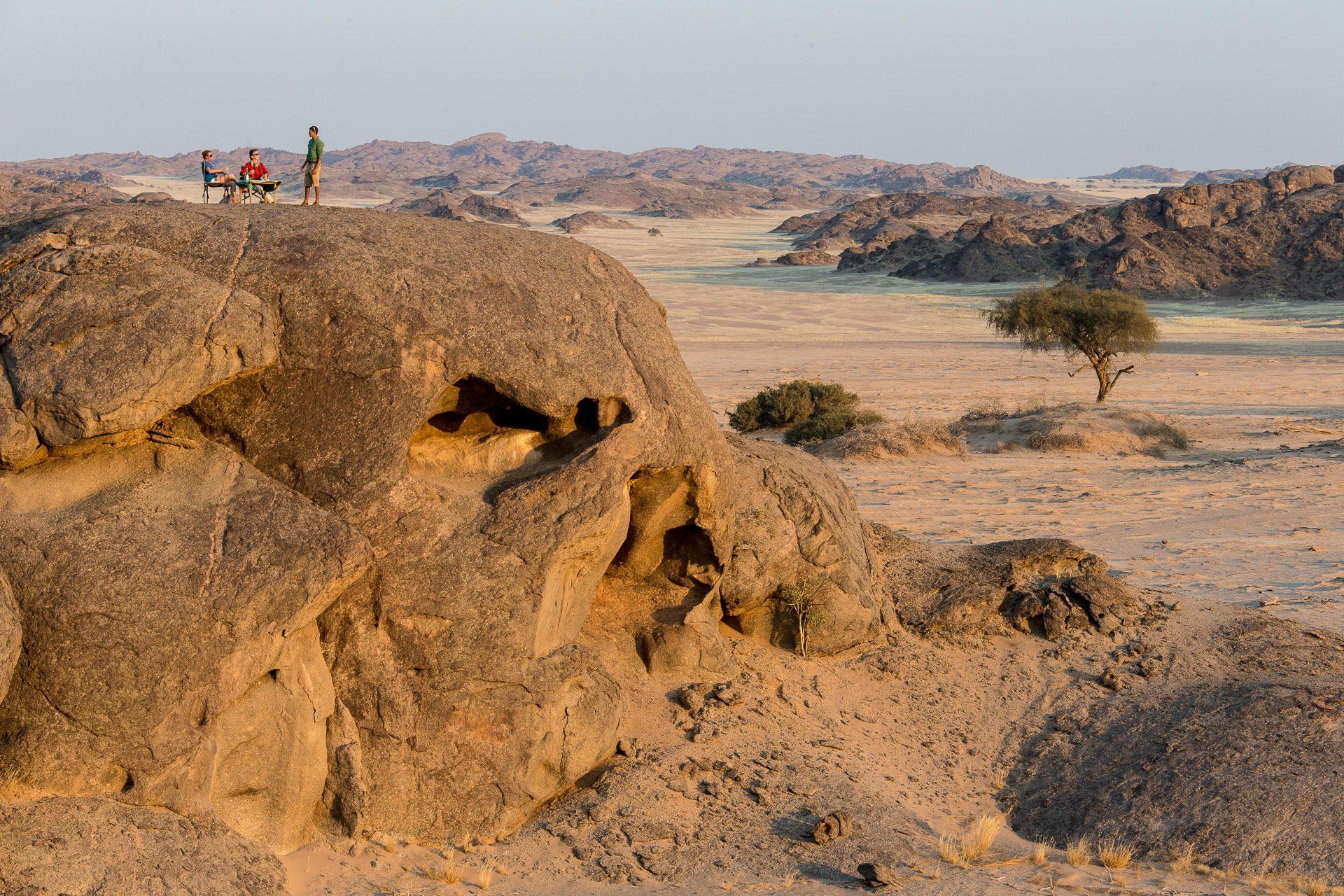 Three people stand atop a large, craggy desert boulder overlooking a vast, arid landscape with sparse vegetation.