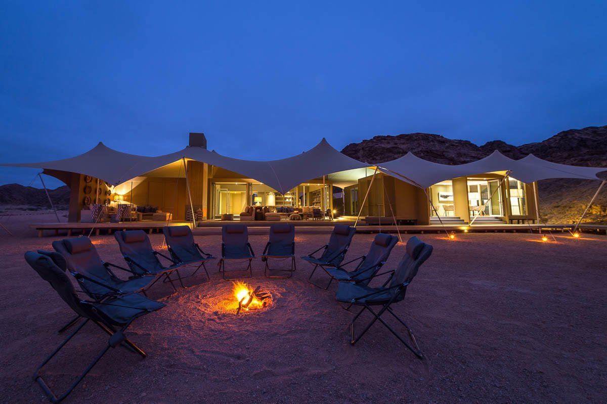 A campfire glows in front of an open, tented desert lodge under a twilight sky.