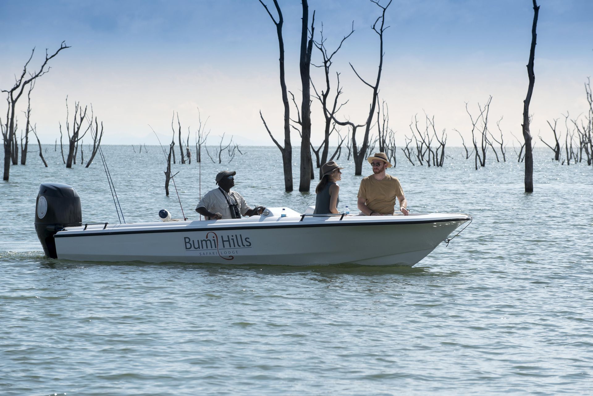 Three people travel on a white motorboat through calm water filled with bare, vertical tree trunks under a cloudy sky.