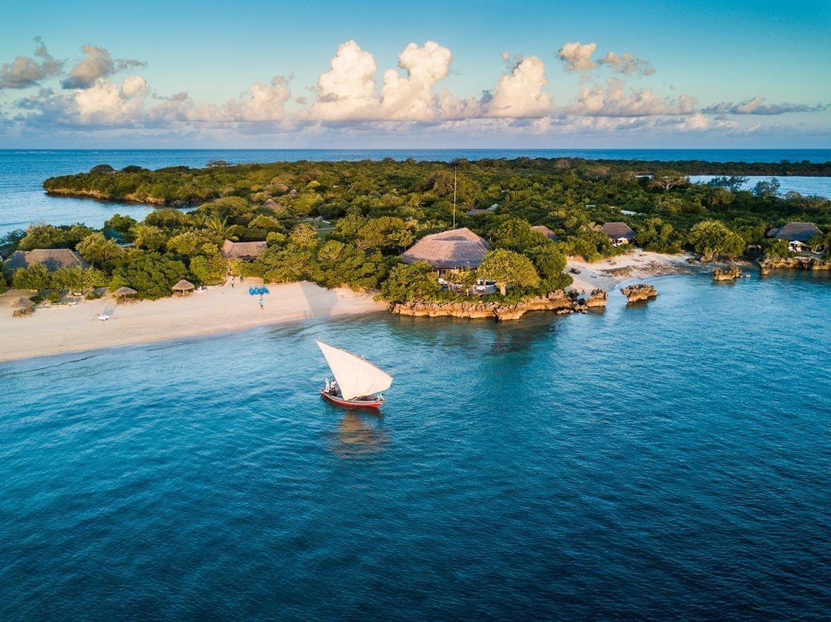A traditional wooden sailboat with a white triangular sail floats on turquoise water near a lush tropical island beach.