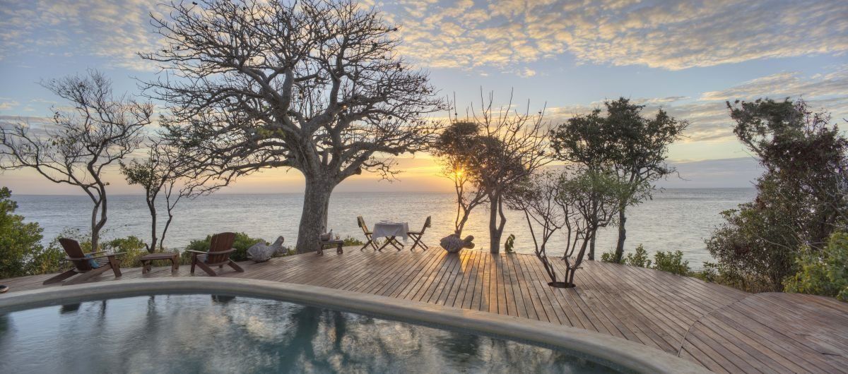 A pool deck with trees and chairs overlooking a calm sea at sunset.