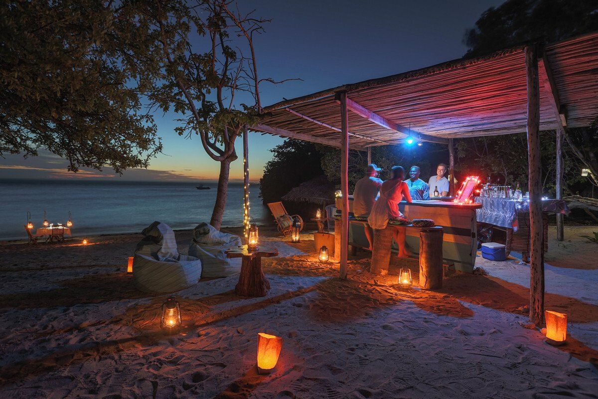 People gather at a candlelit beach bar under a thatched roof during twilight, with soft glowing lights along the sand.
