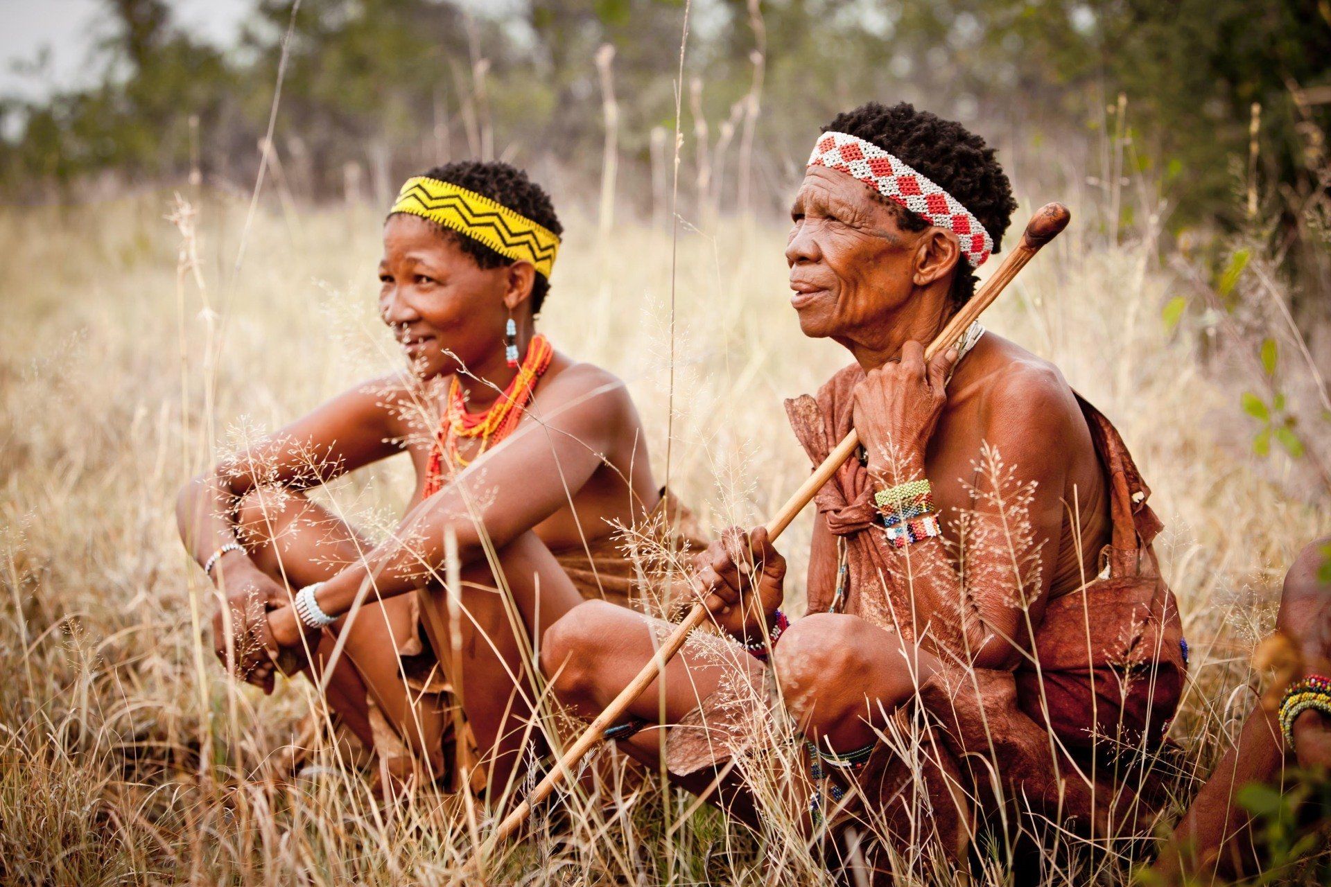 Two people wearing beaded headbands and traditional clothing sit in a tall, dry grass field, one holding a wooden staff.