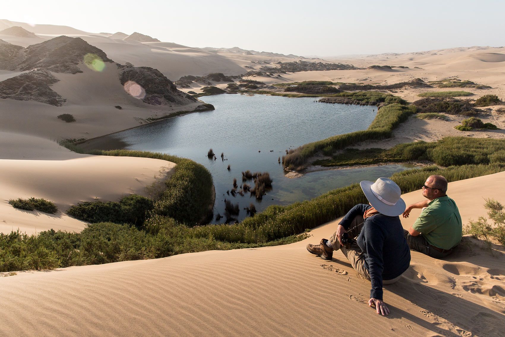 Two people sit on a sand dune looking out over a desert oasis surrounded by lush green vegetation.