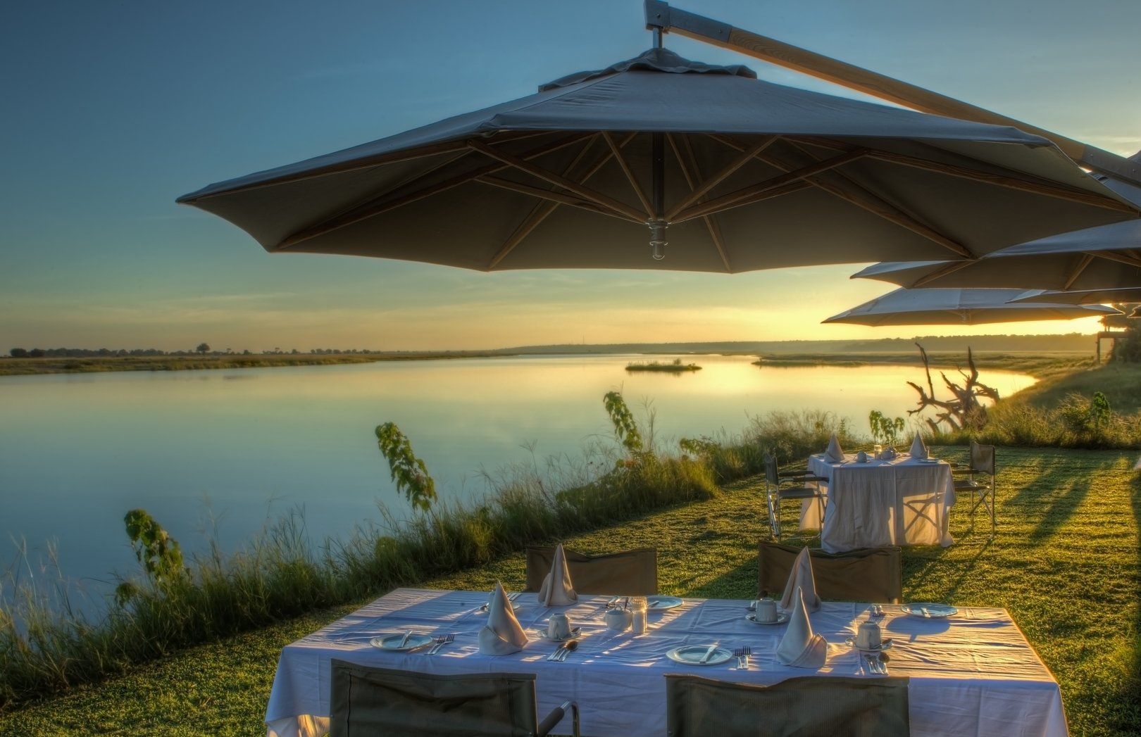 Tables set for outdoor dining under large umbrellas at sunset, overlooking a calm riverbank.