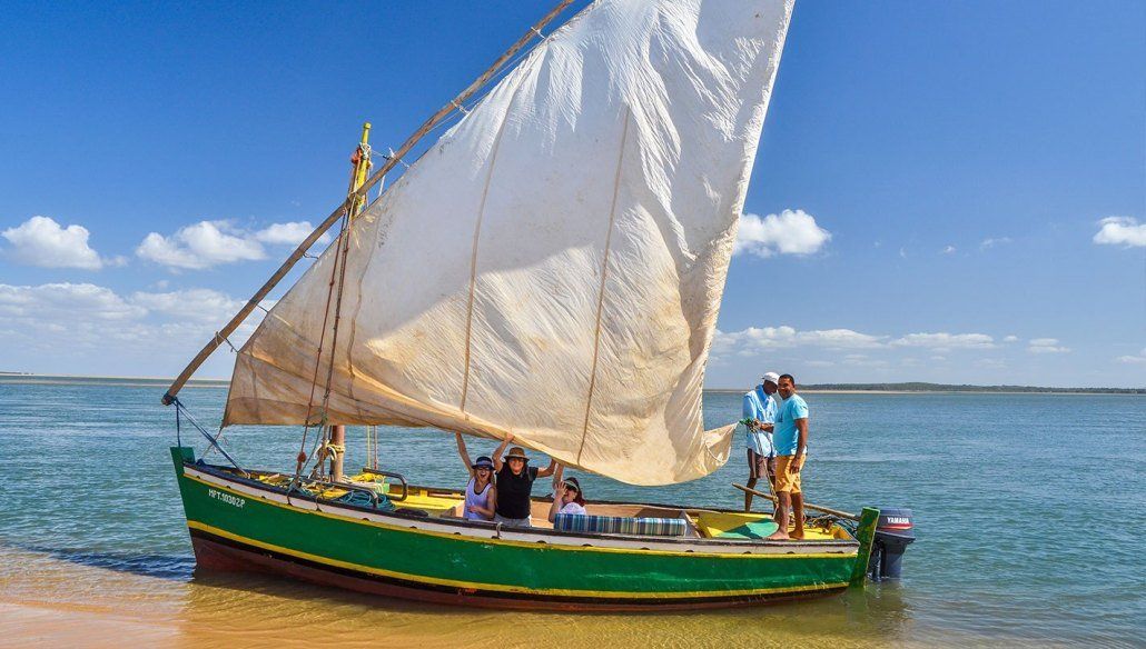 A small, green sailboat with a white triangular sail sits in calm, shallow water under a blue sky with scattered clouds.