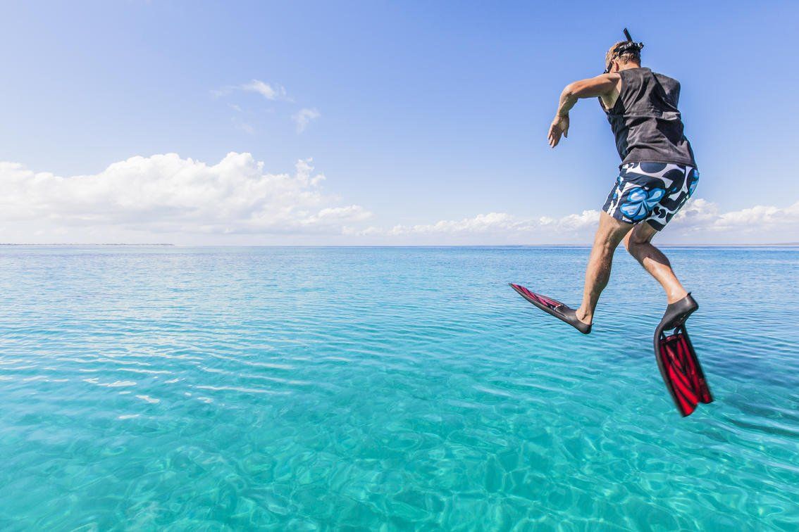 A person wearing a life vest and flippers leaps into clear turquoise tropical water under a bright blue sky.