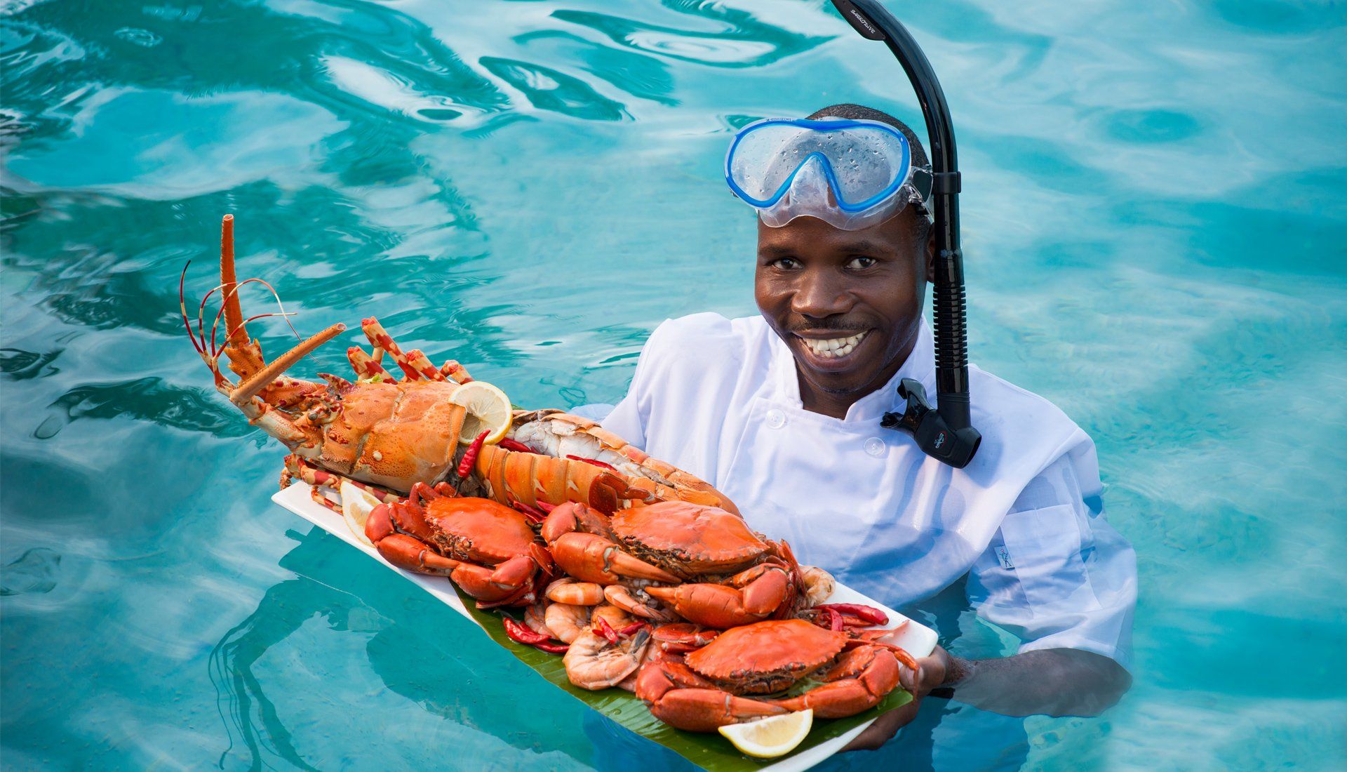 A smiling person in a chef's uniform and snorkel gear holds a large platter of cooked seafood in bright blue water.