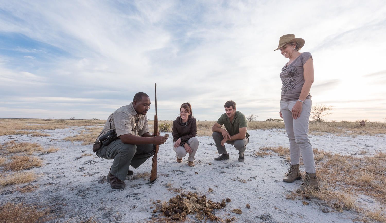 A guide holds a rifle and shows animal tracks to a group in a sunny, dry savanna landscape.