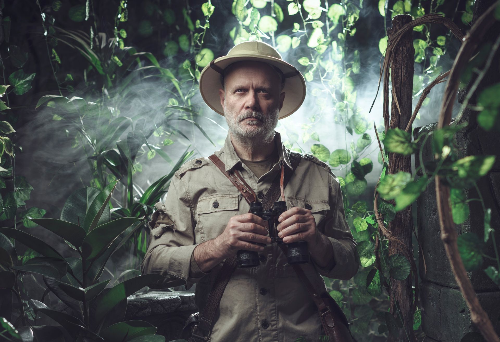 A person in a pith helmet and safari shirt holds binoculars in a misty, lush jungle setting.