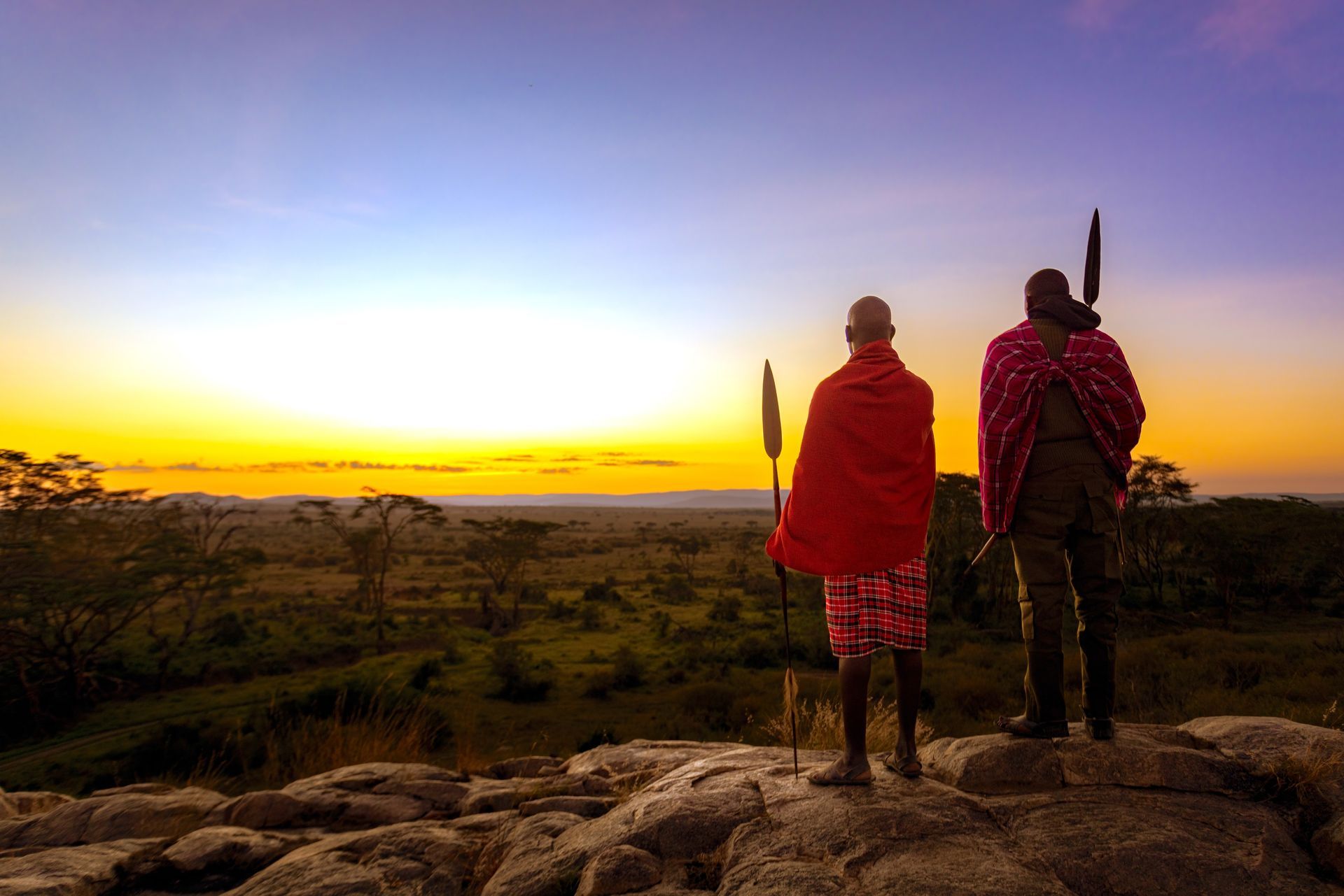 Two people in traditional shuka wraps stand on rocky terrain, looking over a vast savanna during a golden sunset.