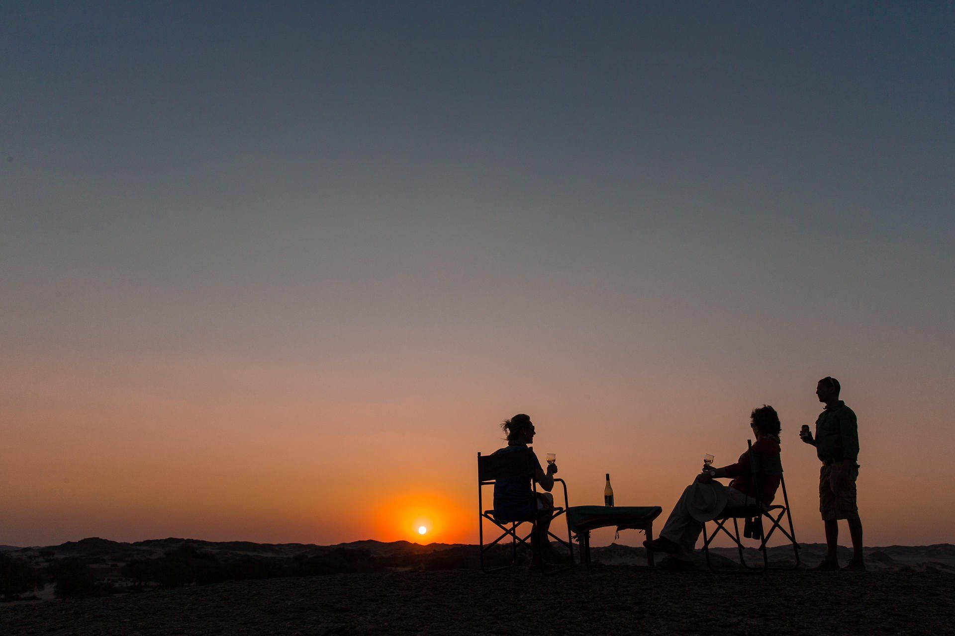 Silhouettes of three people enjoying drinks at a small table while watching the sunset over a flat, rocky landscape.