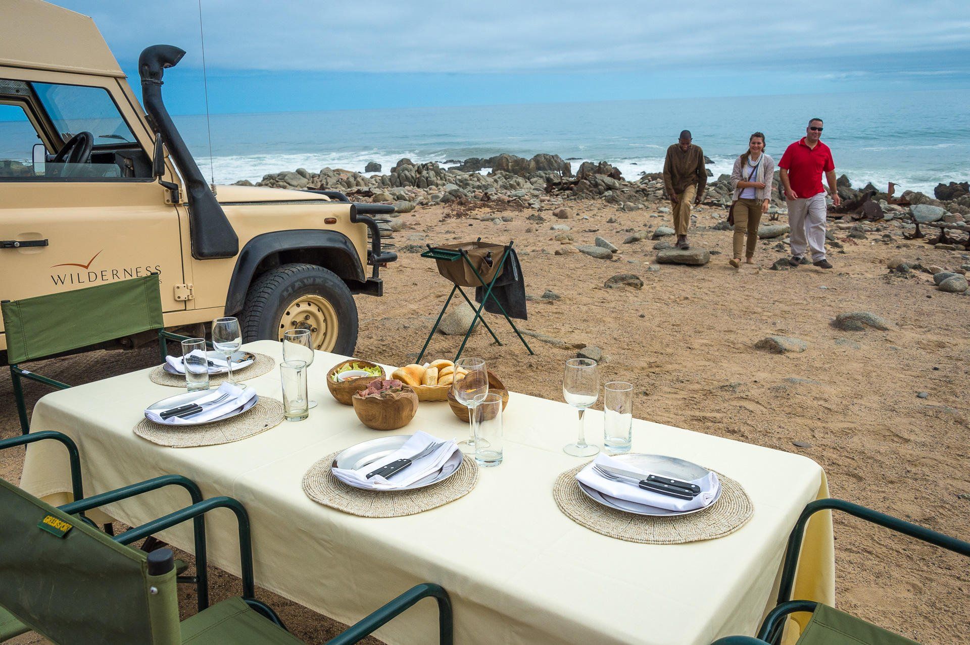 A dining table set for four on a rocky beach next to a parked safari vehicle, with three people standing in the distance.