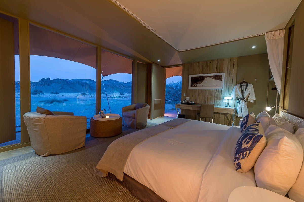 A luxury tented bedroom featuring a large bed, seating area, and panoramic floor-to-ceiling windows overlooking nature.