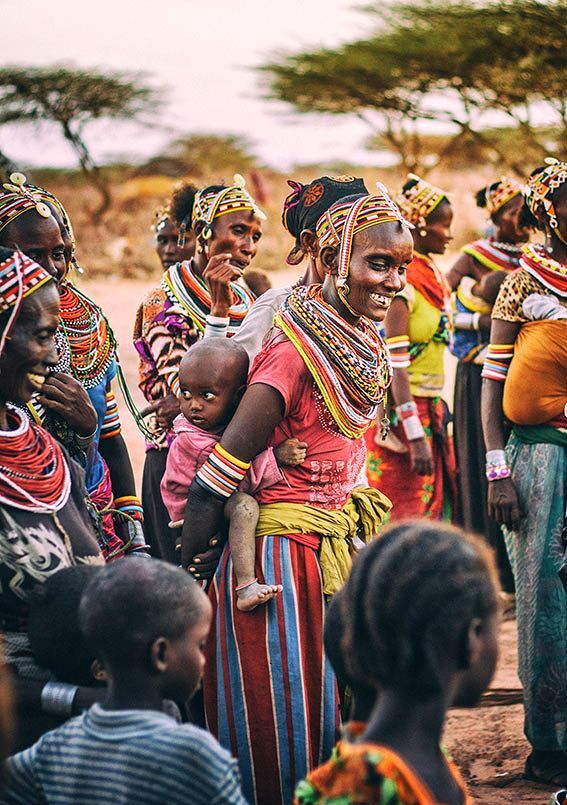 A group of people wearing intricate beaded necklaces and colorful patterned clothing in a dry, outdoor landscape.
