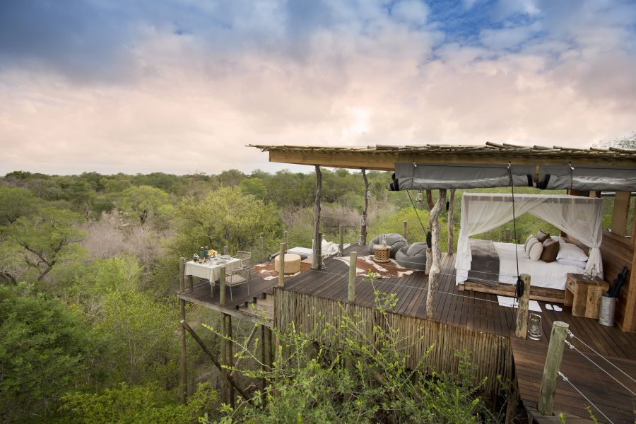 An elevated open-air wooden deck with a bed, dining table, and seating, overlooking a dense, lush green forest.
