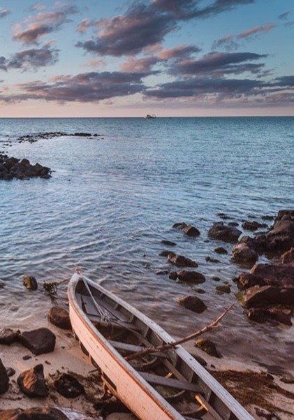 A weathered rowboat rests on a rocky shore beside calm ocean waters under a cloudy sunset sky.