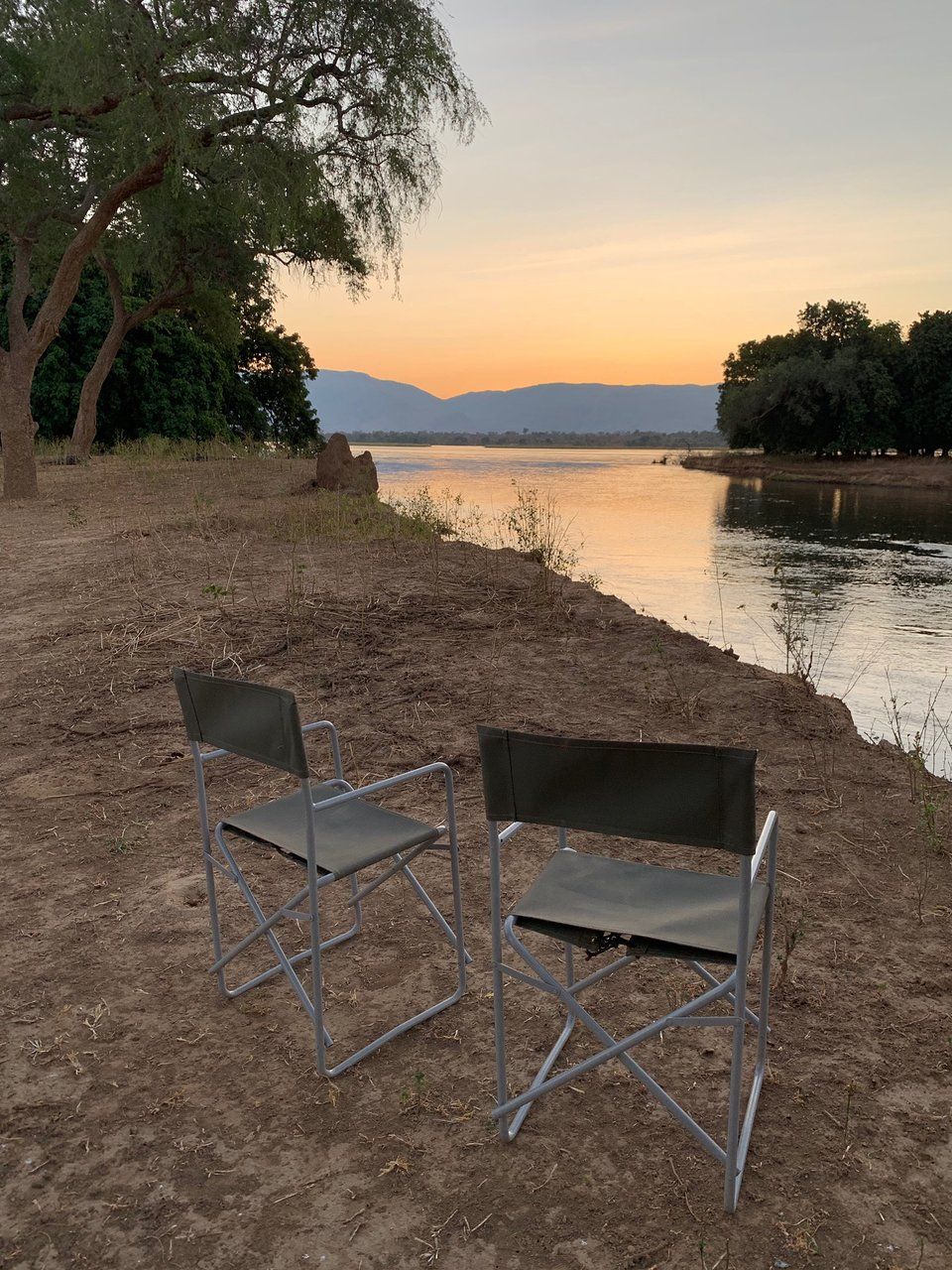 Two folding chairs sit on a dry, dirt riverbank at sunset, facing a calm body of water with mountains in the distance.