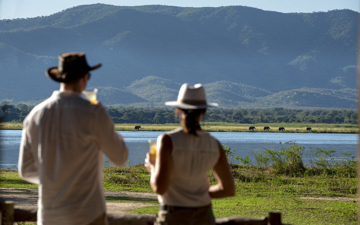 Two people wearing sun hats stand overlooking a river with elephants in the distance against a backdrop of mountains.
