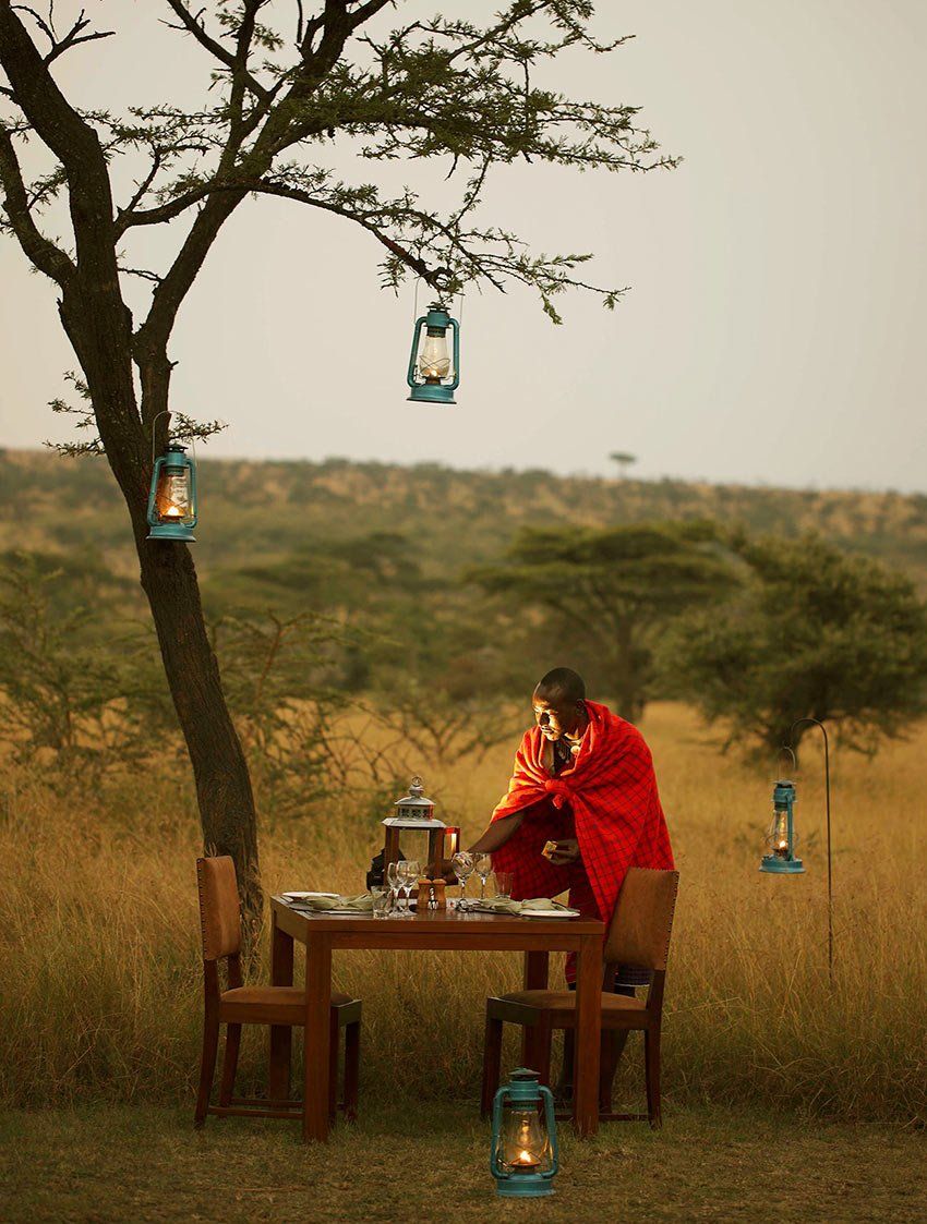 A person in a red garment sets a table outdoors in a grassy savanna at twilight, illuminated by hanging lanterns.