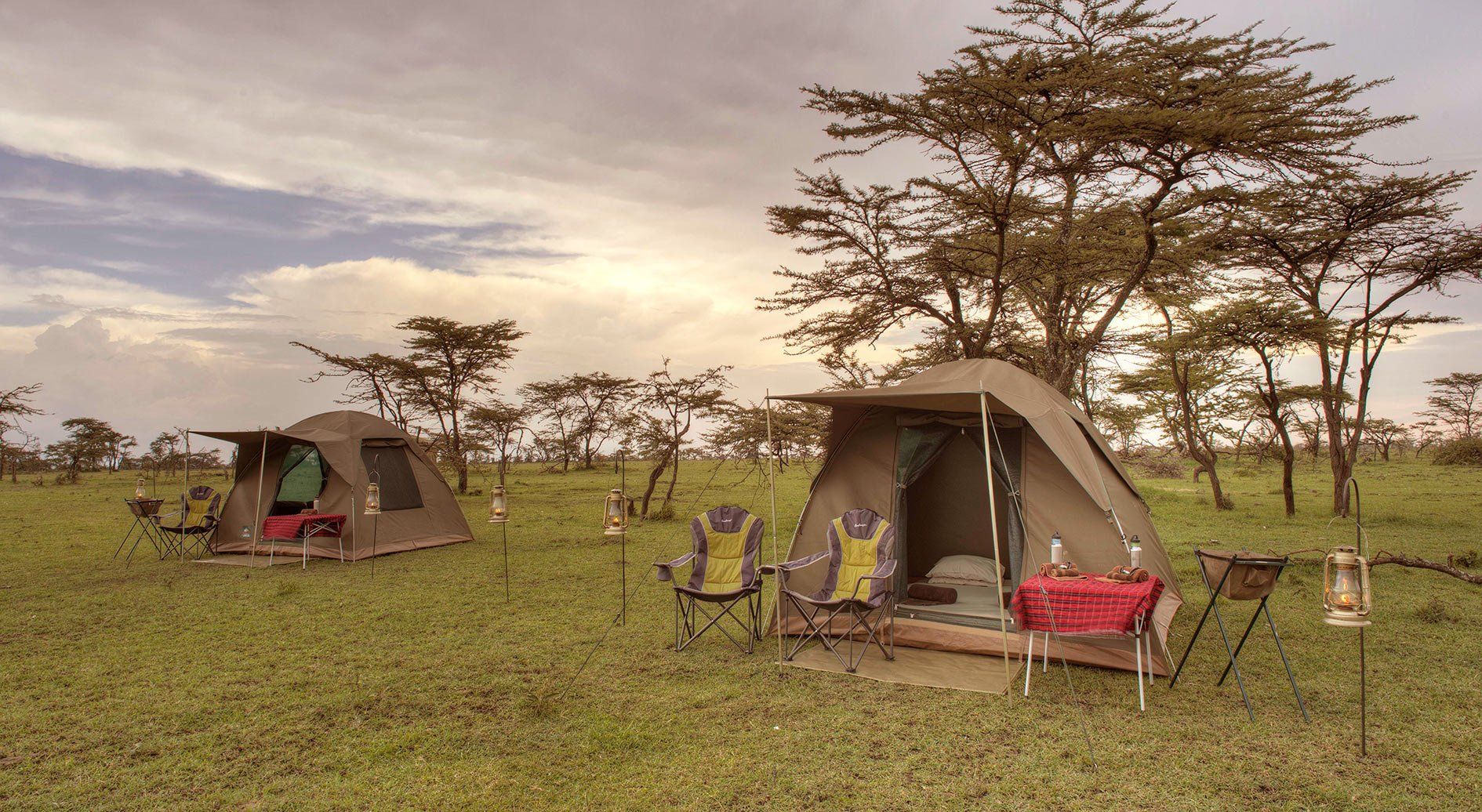 Two safari tents set up in a grassy savanna landscape with scattered acacia trees under a cloudy sunset sky.