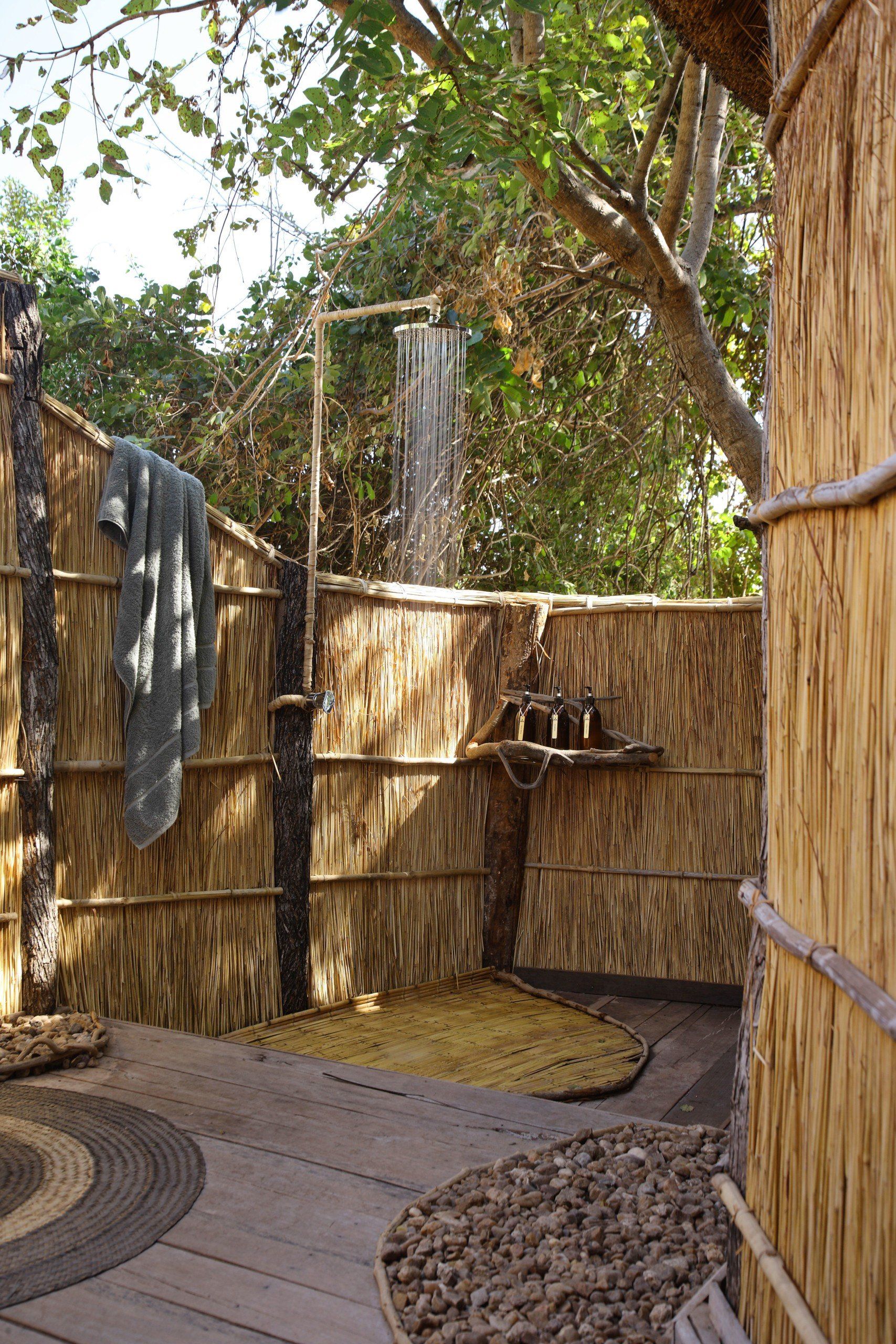 An outdoor shower in a natural setting, featuring a rustic thatched wall, wooden floor, and a towel hanging on a post.