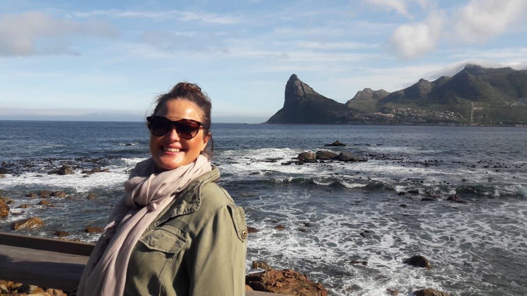 A smiling person wearing sunglasses and a scarf stands by the sea, with a prominent, jagged mountain peak in the distance.