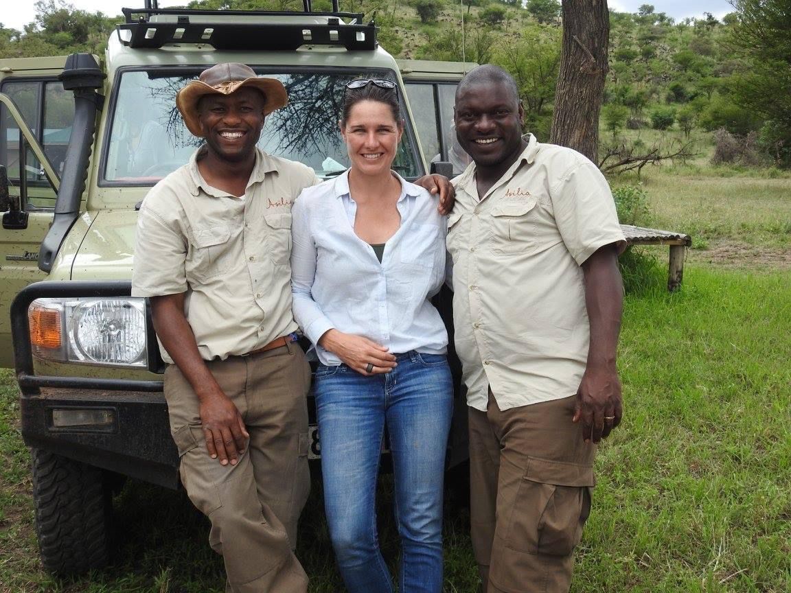 Three people stand smiling in front of a parked safari vehicle in a grassy, outdoor setting.