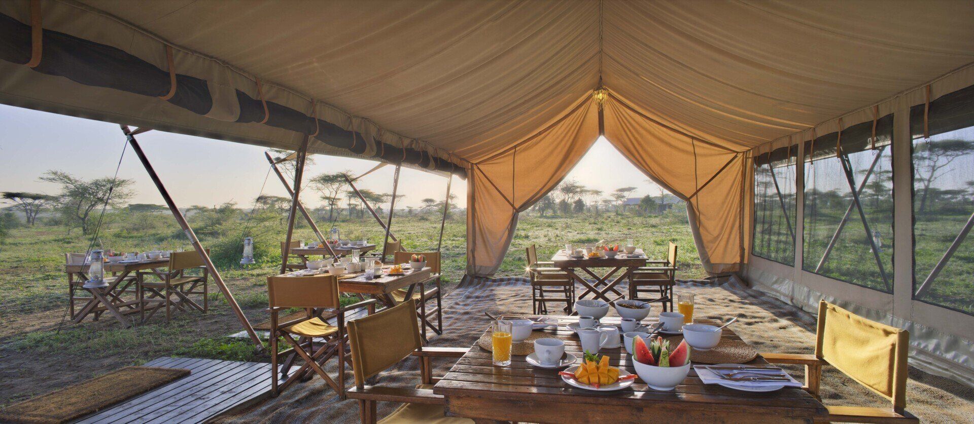 A dining area under an open-sided safari tent with multiple tables and chairs set in a grassy landscape at sunrise.