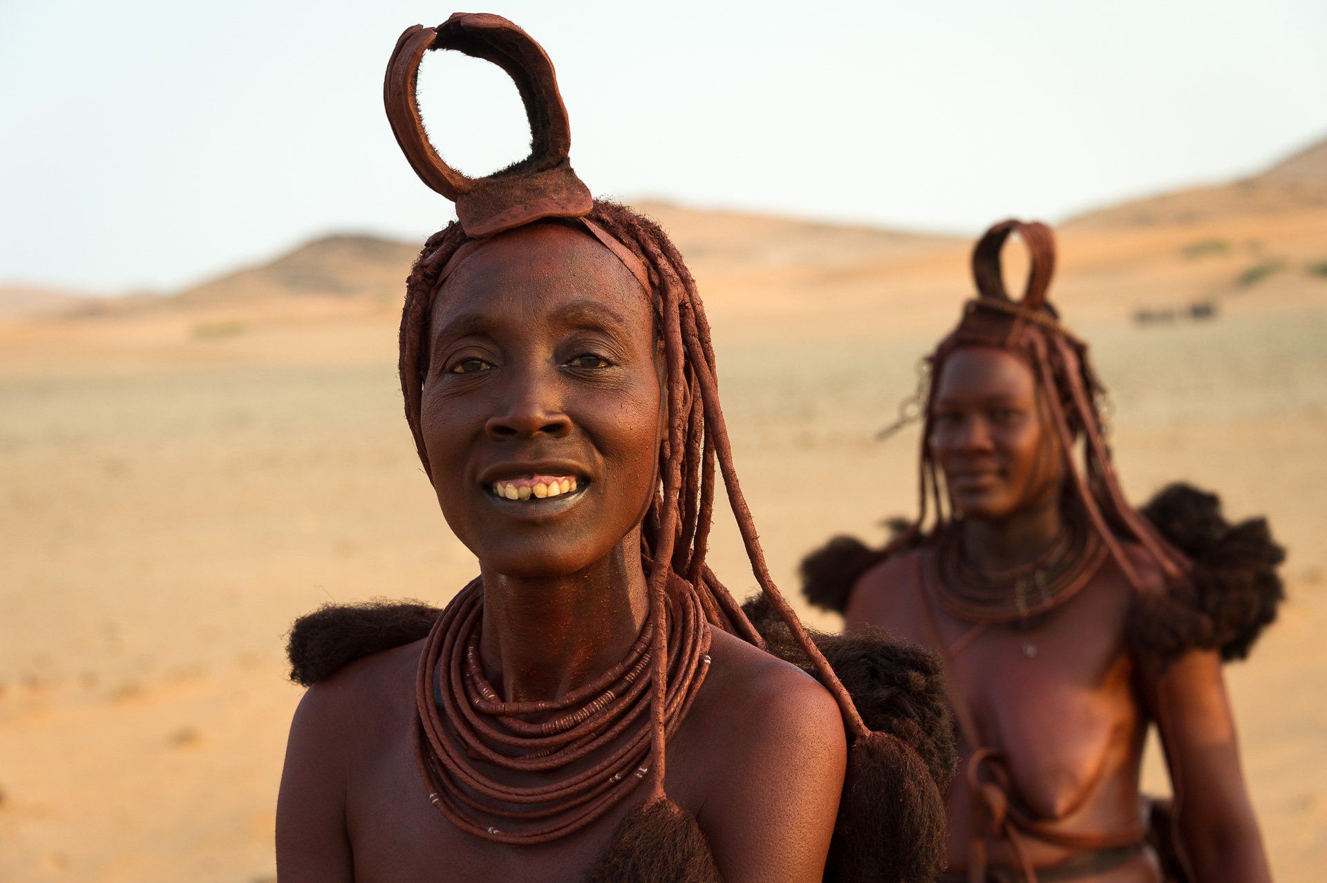 Two Himba women with traditional ochre-covered hair and skin, adorned in beaded necklaces, standing in a desert landscape.