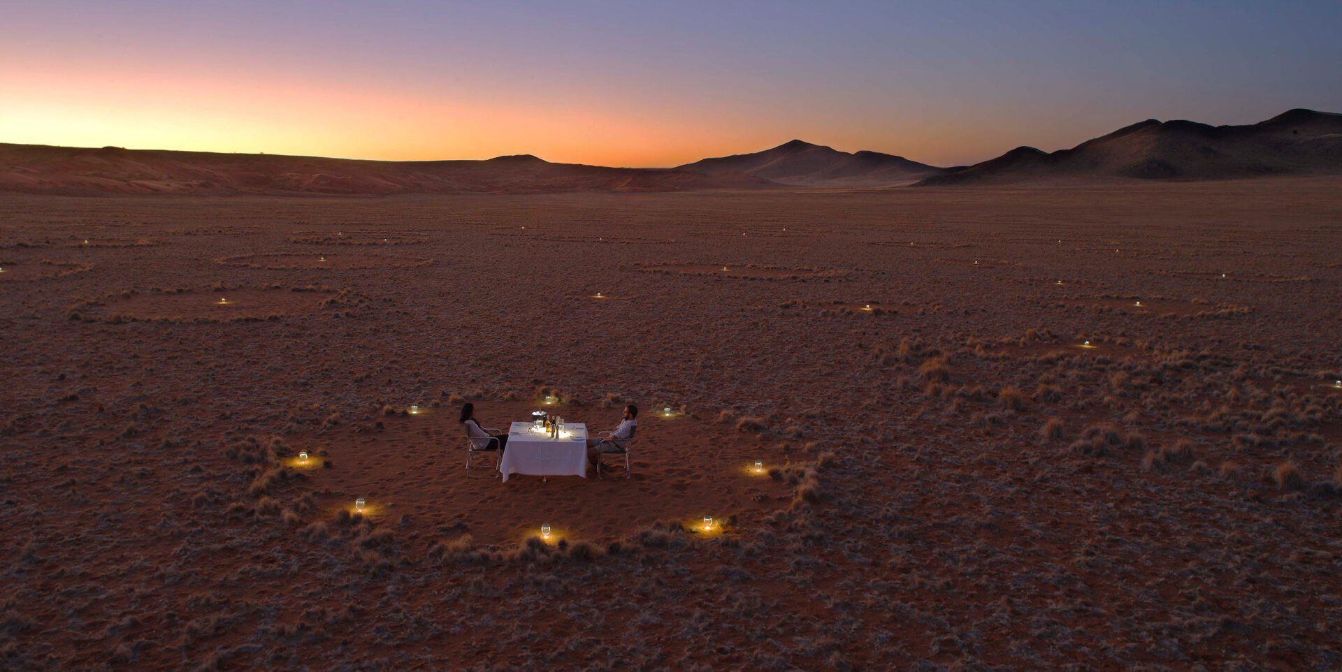 Two people dine at a small table surrounded by a circle of glowing lanterns in a vast, arid desert at sunset.