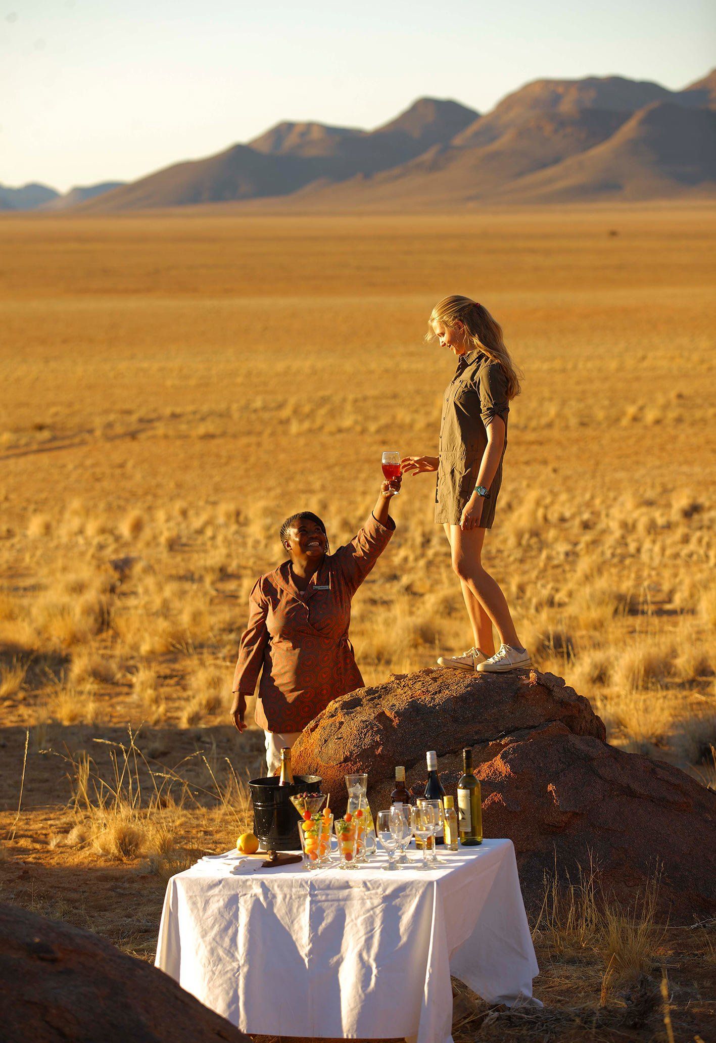 Two people toast with drinks on a rocky outcrop in a vast, golden desert landscape, beside a small table with wine glasses.