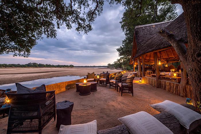 Patio seating at a safari lodge at twilight, overlooking a dry riverbed under a cloudy sky with warm ambient lighting.