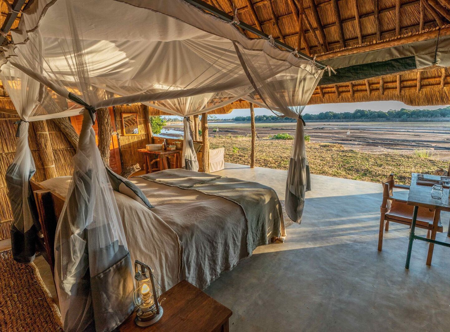 An open-air safari lodge bedroom featuring a canopy bed, straw roof, and a scenic view of a dry riverbed.