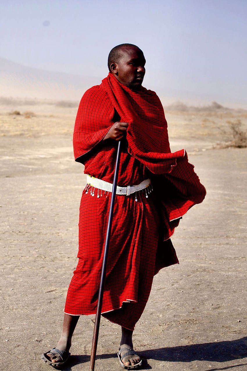 A person wearing a bright red shuka cloth and sandals stands in a desert landscape, leaning on a tall walking stick.