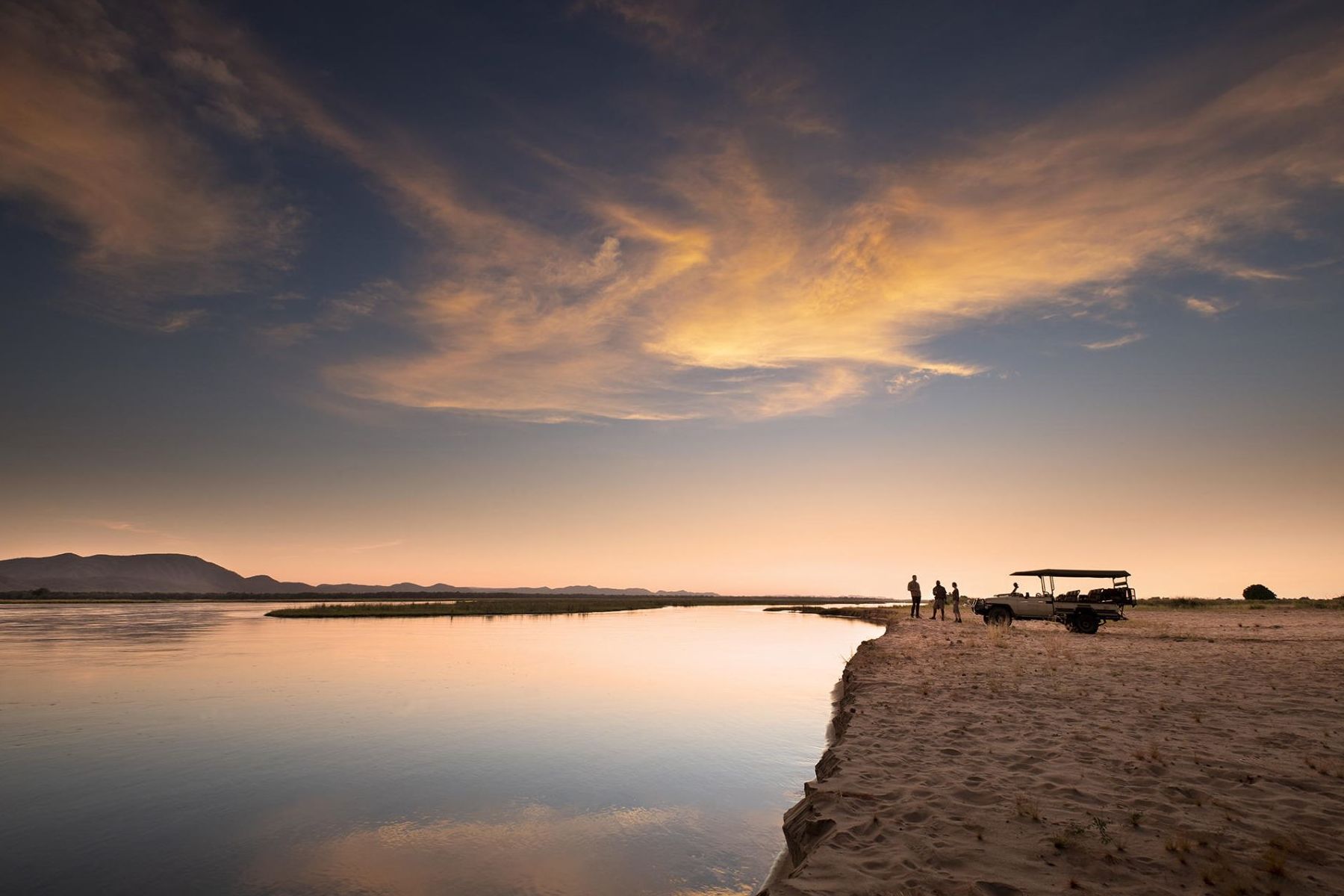 A safari vehicle and people on a sandy riverbank at sunset, with golden clouds reflected in the calm, wide river.