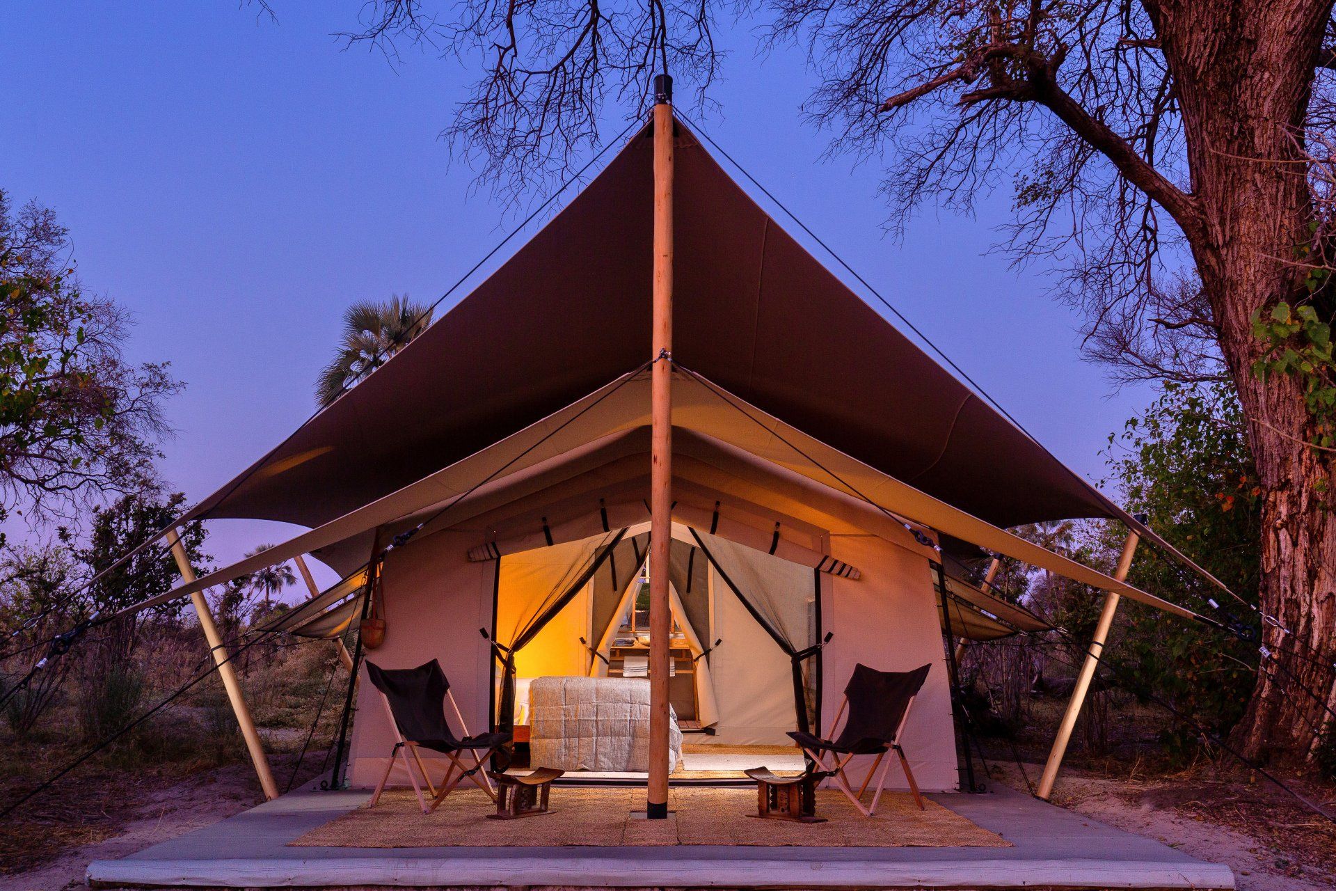 A luxury safari tent at dusk with an open front entrance, two folding chairs on a deck, and an illuminated interior.