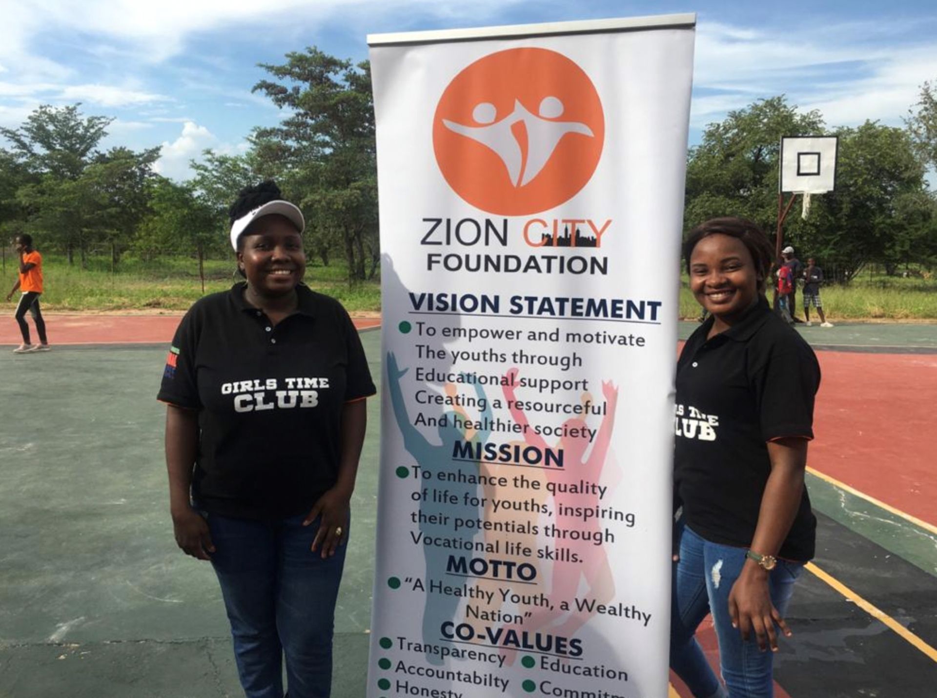 Two smiling individuals stand on a basketball court beside a Zion City Foundation banner detailing its mission and values.