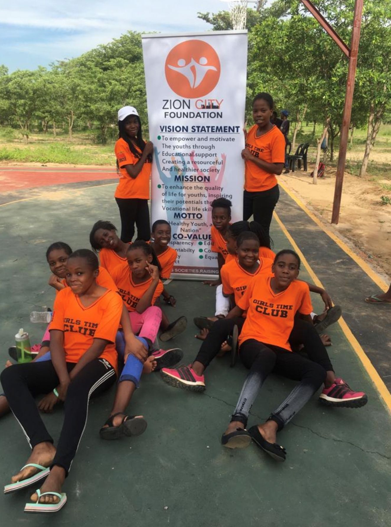 A group of people wearing orange shirts gather around a Zion Foundation banner outdoors on a sports court.