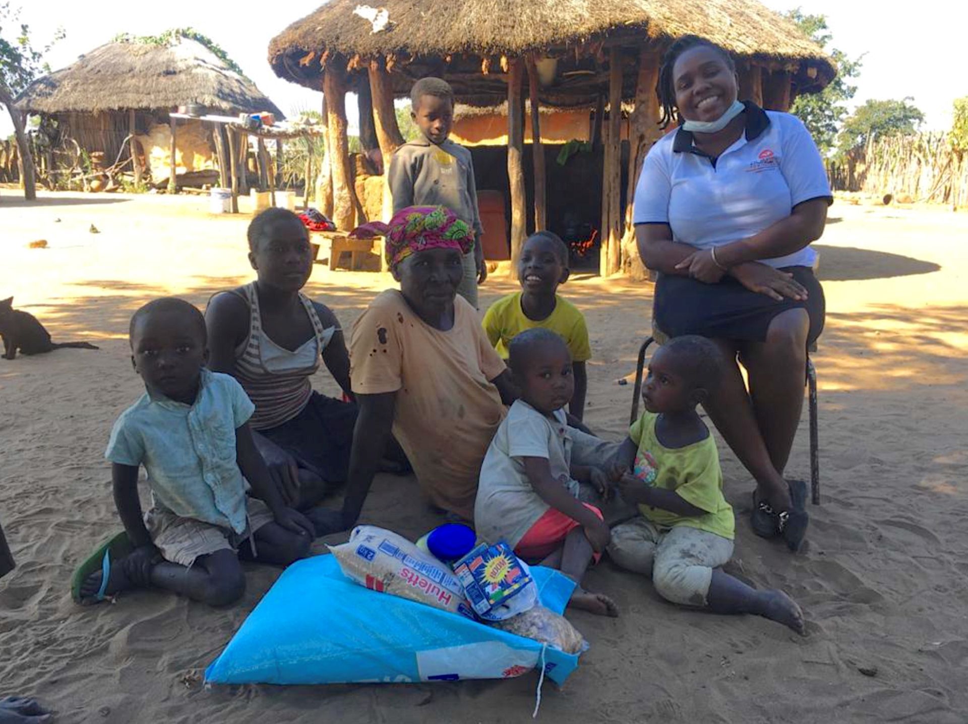 A group sits in a sandy rural yard with traditional huts. A person in a white polo shirt poses with children and goods.