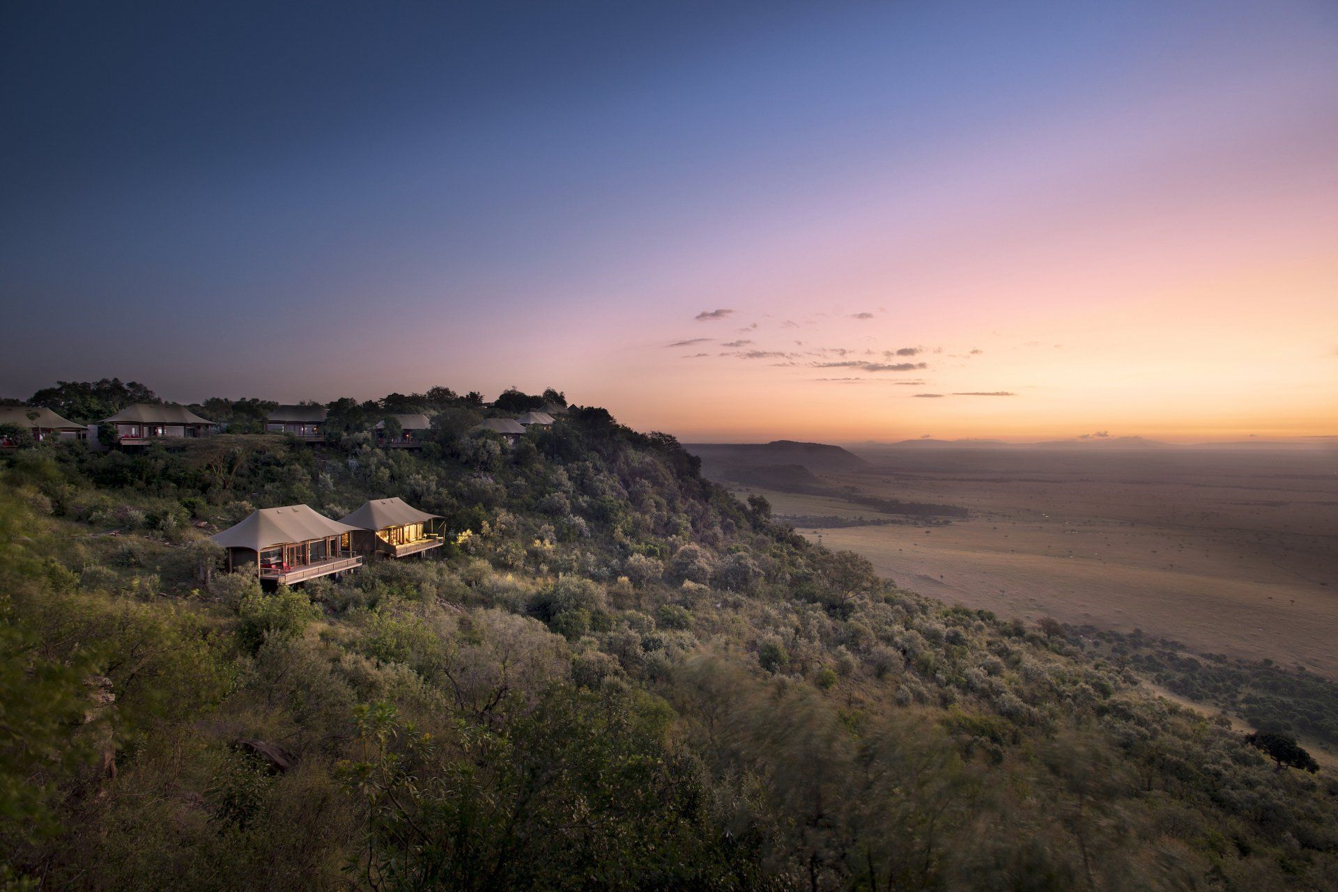 Tented safari lodges perched on a green, rocky hillside at sunset, overlooking an expansive, hazy savanna landscape.
