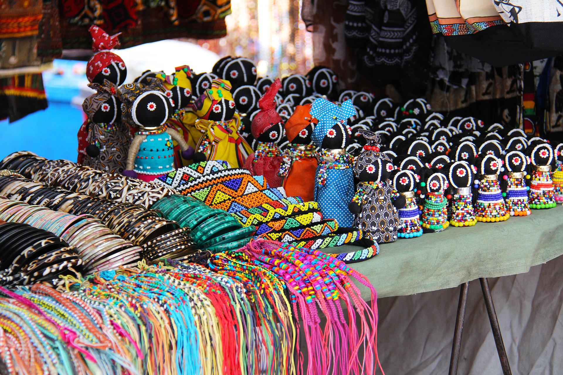 A market stall displays colorful beaded dolls, intricate bracelets, and rows of vibrant thread necklaces on a table.