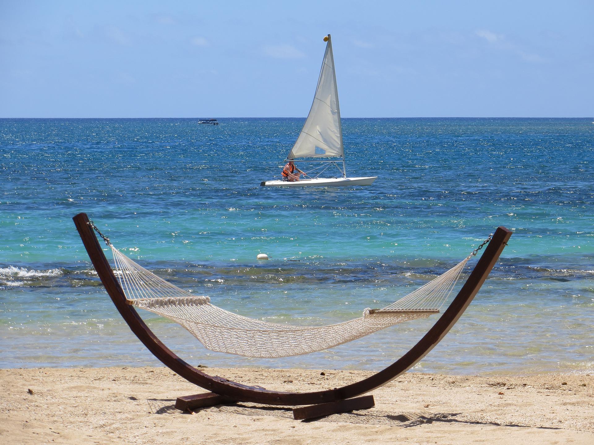 A white hammock stands on a sandy beach, framing a small sailboat drifting on the turquoise ocean under a clear blue sky.