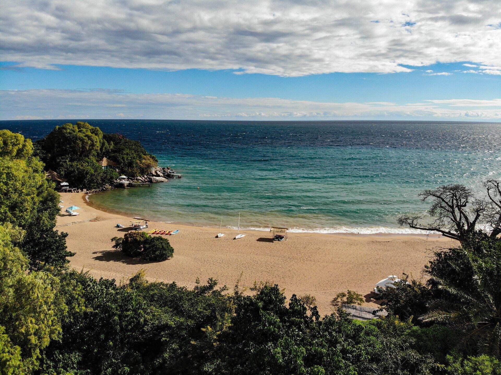 A sandy beach cove surrounded by lush trees, meeting the blue water of a vast lake under a partly cloudy sky.