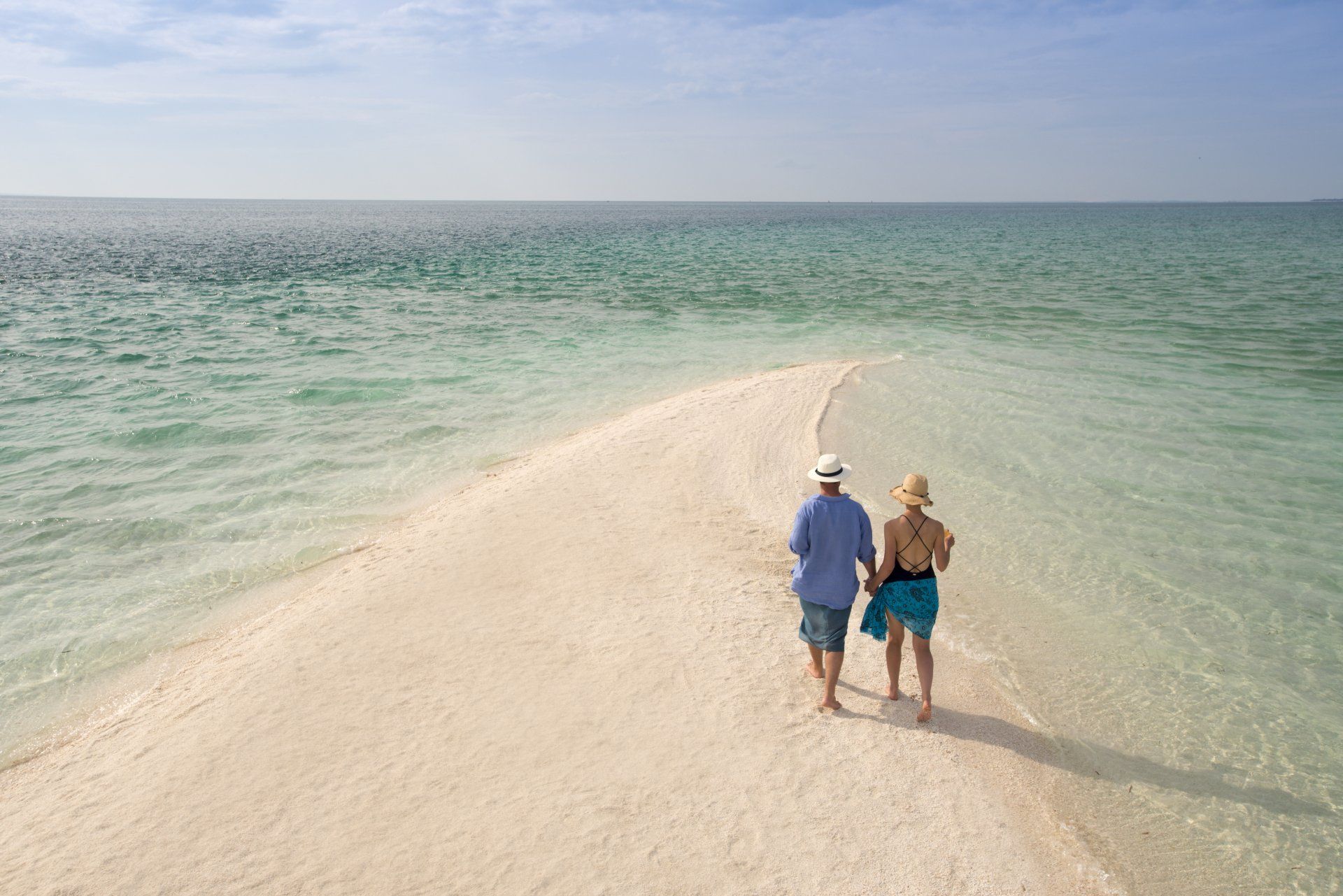 Two people in hats walk along a narrow, sandy white beach surrounded by calm, clear turquoise ocean water.